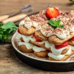 Zu sehen ist ein Holztisch, auf dem ein weisser Teller steht, auf dem eine Portion Erdbeer-Tiramisu angerichtet ist. Daneben liegt Besteck mit Holzgriffen. [Foto: AdobeStock_Надія Коваль]