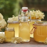 Holunderblütensirup in Gläsern und Flaschen abgefüllt. Hier ist im Hintergrund ein Garten zu sehen. Zwischen dem Sirup liegen Holunderblüten. [Foto: AdobeStock_Madeleine Steinbach]