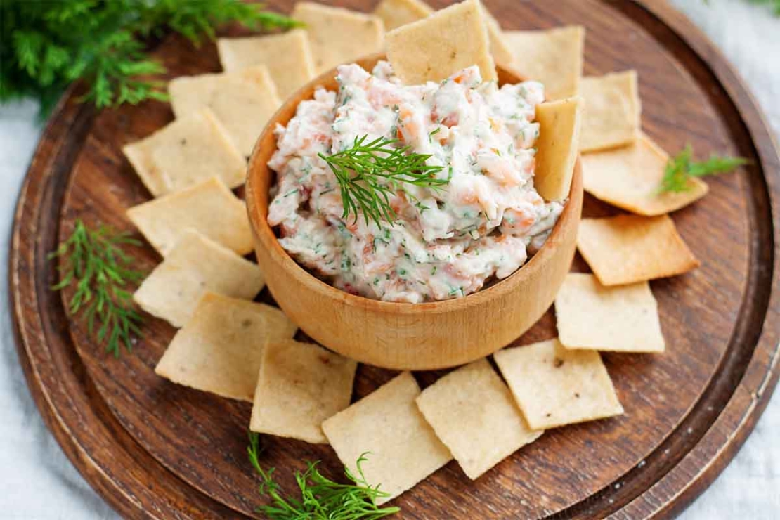 Schale mit Lachs-Creme auf einer Unterlage aus dunklem Holz. Die Creme oder der Dip wird hier serviert mit Crackern, die kreisfoermig um den Dip ausgelegt wurden. Auf dem Dip sind Dill-Blaetter zu sehen.[Foto: AdobeStock_filirochka]