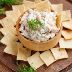 Schale mit Lachs-Creme auf einer Unterlage aus dunklem Holz. Die Creme oder der Dip wird hier serviert mit Crackern, die kreisfoermig um den Dip ausgelegt wurden. Auf dem Dip sind Dill-Blaetter zu sehen.[Foto: AdobeStock_filirochka]