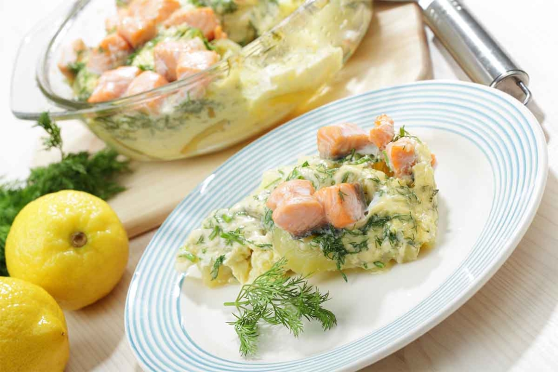 Auf dem Bild ist Lachs aus dem Ofen in einer Auflaufform aus Glas zu sehen, der hinter einem weissen Teller und neben einem Paar Zitronen auf einem Tisch steht. Der Ofenlachs wurde in einer hellen Cremesauce mit Dill und Kartoffeln gebacken. Ein Teil aus der Auflaufform ist auf dem weissen Teller angerichtet. [Foto: AdobeStock_StockphotoVideo]