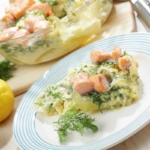 Auf dem Bild ist Lachs aus dem Ofen in einer Auflaufform aus Glas zu sehen, der hinter einem weissen Teller und neben einem Paar Zitronen auf einem Tisch steht. Der Ofenlachs wurde in einer hellen Cremesauce mit Dill und Kartoffeln gebacken. Ein Teil aus der Auflaufform ist auf dem weissen Teller angerichtet. [Foto: AdobeStock_StockphotoVideo]