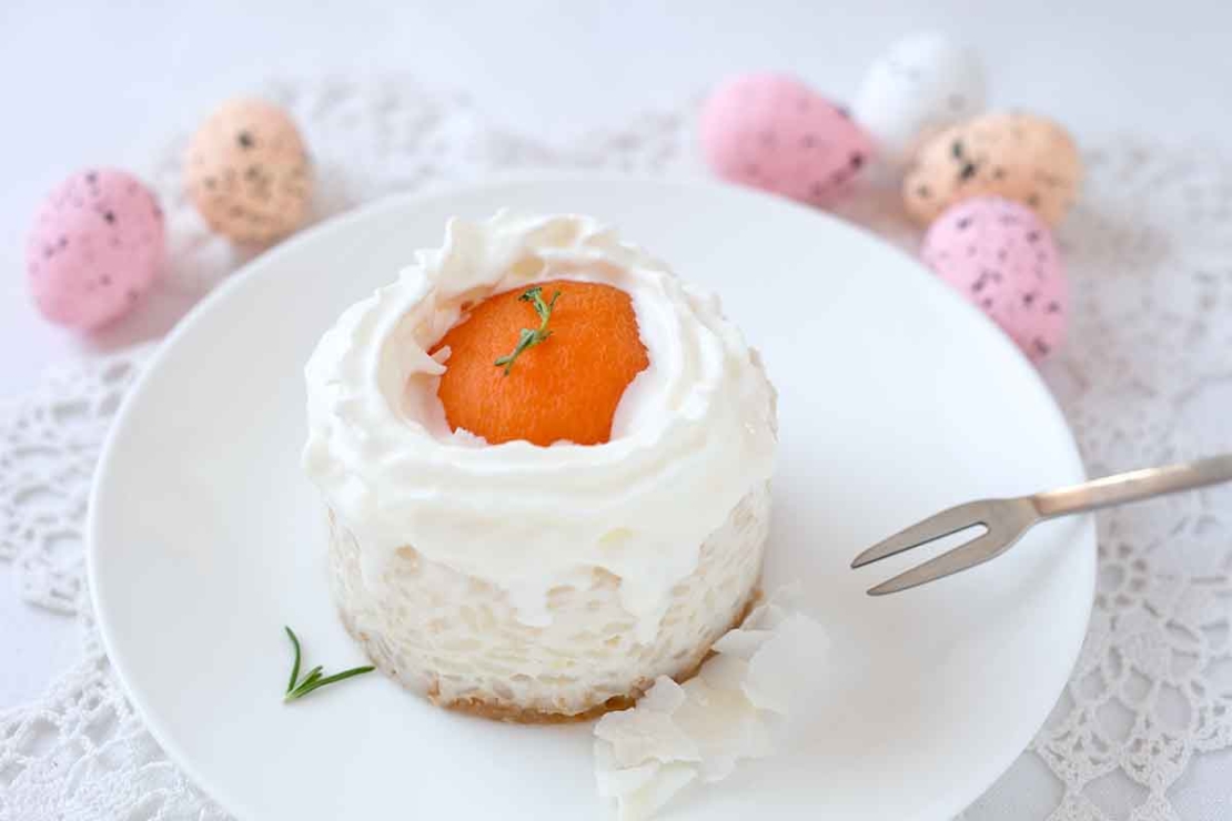 Oster-Toertchen mit Apriokose und Milchreis. Dieses Rezept zu Ostern wird auf einem weissen Teller auf einem weissen Tisch mit bunten Ostereiern im Hintergrund serviert. Das Toertchen ist von der Sahne und dem Milchreis sehr weiss und hat eine halbe Dosenaprikose auf dem oberen Rand, um die ein Kreis aus Schlagsahne gespritzt wurde. [Foto: AdobeStock_rainbow33]