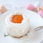 Oster-Toertchen mit Apriokose und Milchreis. Dieses Rezept zu Ostern wird auf einem weissen Teller auf einem weissen Tisch mit bunten Ostereiern im Hintergrund serviert. Das Toertchen ist von der Sahne und dem Milchreis sehr weiss und hat eine halbe Dosenaprikose auf dem oberen Rand, um die ein Kreis aus Schlagsahne gespritzt wurde. [Foto: AdobeStock_rainbow33]