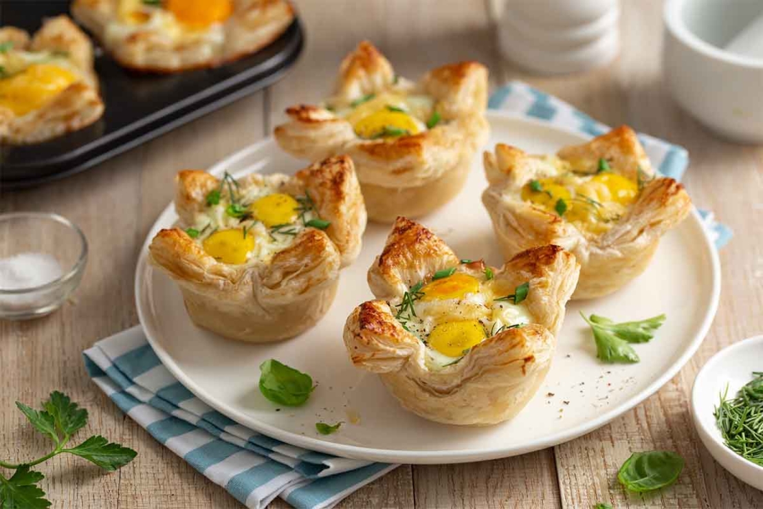 Oster-Muffins aus Blaetterteig mit Ei und Kaese gefuellt. Sie stehen auf einem Tisch auf einem weissen Teller, der auf einem blau-weiss karierten Tuch steht. [Foto: AdobeStock_anna_shepulova]
