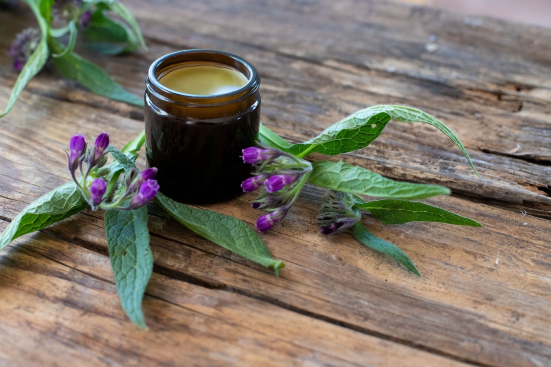 Kleiner Cremetigel aus braunem Glas gefüllt mit einer gelblichen Creme steht auf einem Holztisch, daneben liegen Beinwell-Stängel mit violetten Blüten.