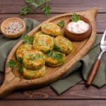 Gemüsefrikadellen liegen auf einem Servier-Tablett aus Holz, auf dem auch ein kleines Schälchen mit Mayonnaise steht. Neben dem Tablett liegen ein Messer, eine Gabel und etwas grober weißer Pfeffer in einem kleinen Schälchen. Das Tablett steht auf einer dunkelgrünen Leinenserviette auf einem Holztisch. [Foto: AdobeStock_Veronika Idiyat]