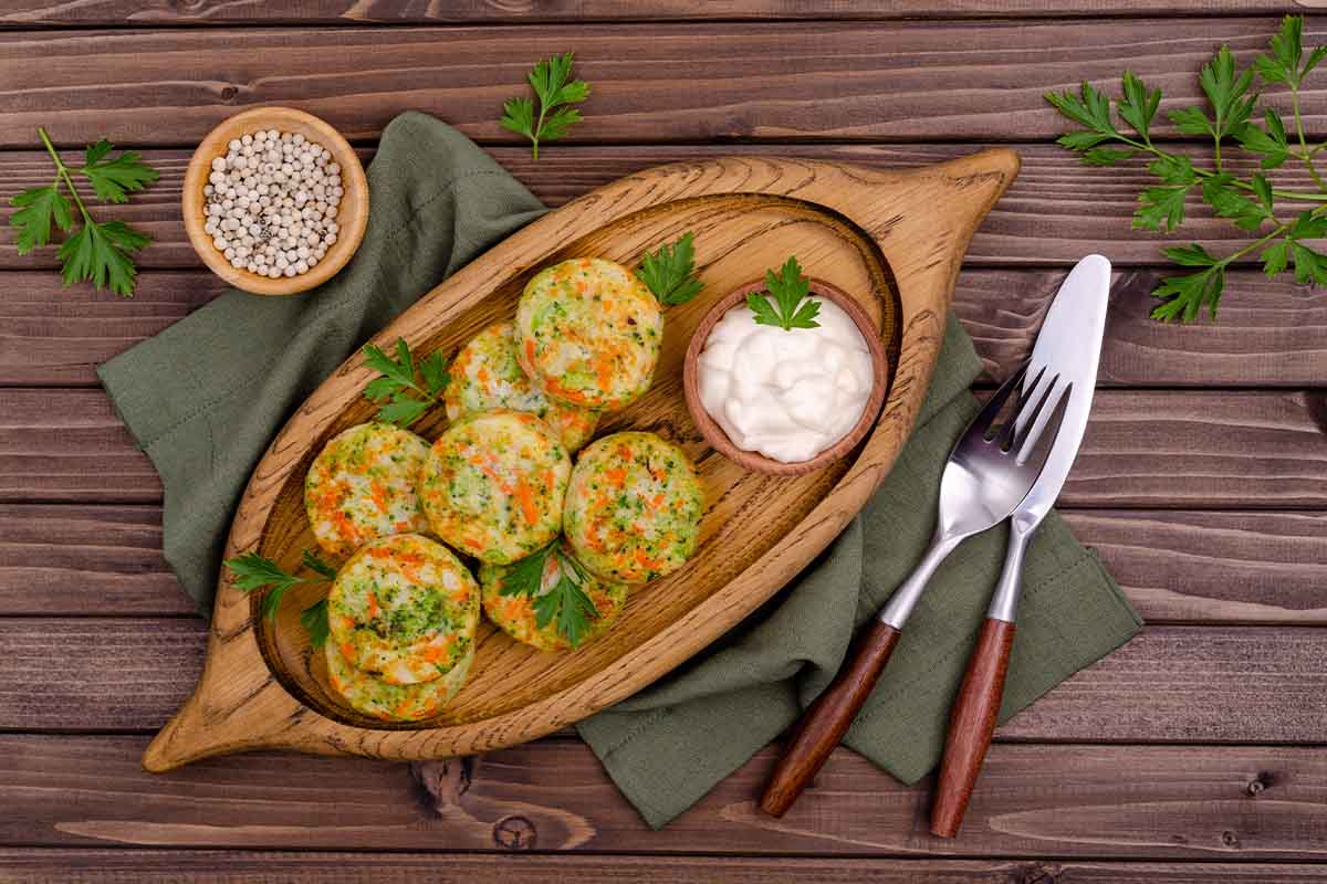 Gemüsefrikadellen liegen auf einem hölzernen Tablett neben einer kleinen Schale Mayonnaise. Rechts daneben befinden sich eine Gabel und ein Messer mit Holzgriff sowie links ein weiteres Schälchen mit weißen Pfefferkörnern. Unter dem Tablett ist eine salbei-grüne Serviette zu sehen. Zudem ist alles mit frischer Petersilie garniert. [Foto: AdobeStock_Veronika Idiyat]