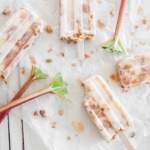 Joghurt-Eis mit Rhabarberstueckchen, das am Stiel gefroren wurde, liegt nebeneinander auf einem weissen Backpapier neben Rhabarber-Stangen auf einem weissen Holztisch. Das Eis ist eine gute Moeglichkeit, eine erfrischende Variante des Stielgemueses zu kreieren, wenn Sie Rhabarber zubereiten moechten. [Foto: AdobeStock_Kati Finell]