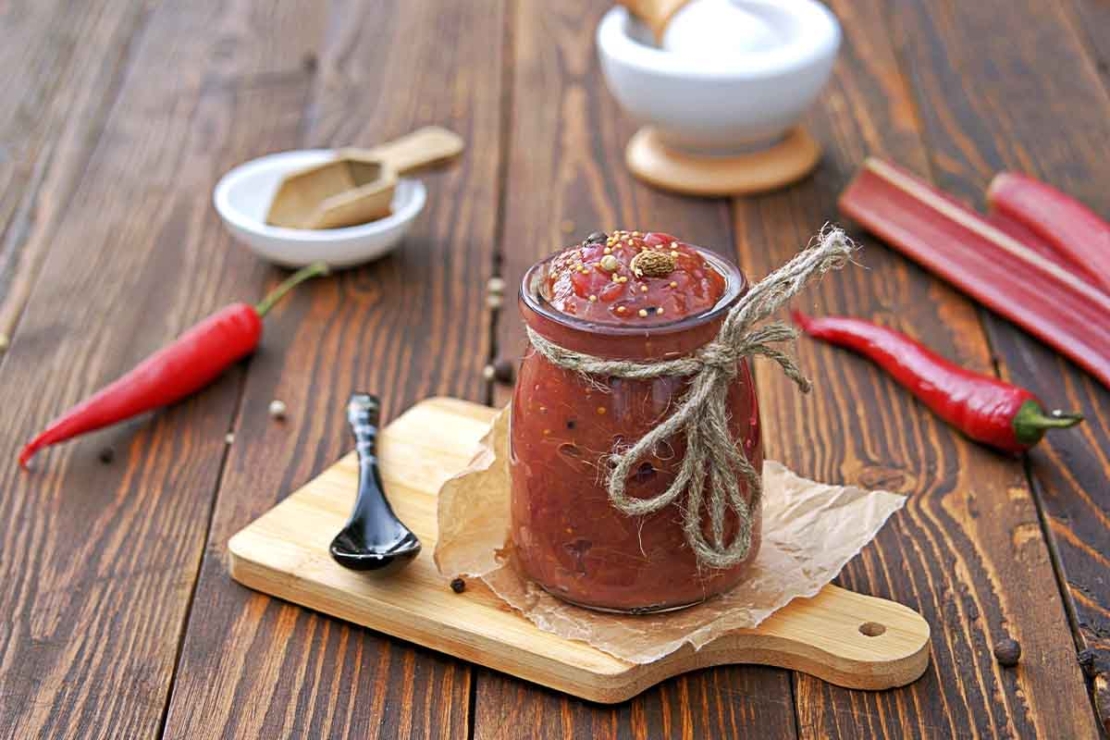 Rhabarber-Sauce oder auch Victoria-Sauce in einem Glas mit einer Schleife auf Juteschnur, das auf einem Holzbrettchen neben einem Loeffel steht. Im Hintergrund sind Gewuerze, Rhabarber und Chilis zu sehen. [Foto: AdobeStock_Natallya_ph]