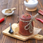 Rhabarber-Sauce oder auch Victoria-Sauce in einem Glas mit einer Schleife auf Juteschnur, das auf einem Holzbrettchen neben einem Loeffel steht. Im Hintergrund sind Gewuerze, Rhabarber und Chilis zu sehen. [Foto: AdobeStock_Natallya_ph]