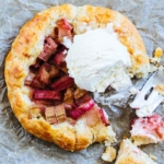 Eine knusprige Galette mit Rhabarberfuellung liegt auf etwas Backpapier auf einem Tisch. Oben auf der mit Rhabarber befuellten Mitte liegt eine Kugel mit leckerem Vanilleeis. [Foto: AdobeStock_Peti]
