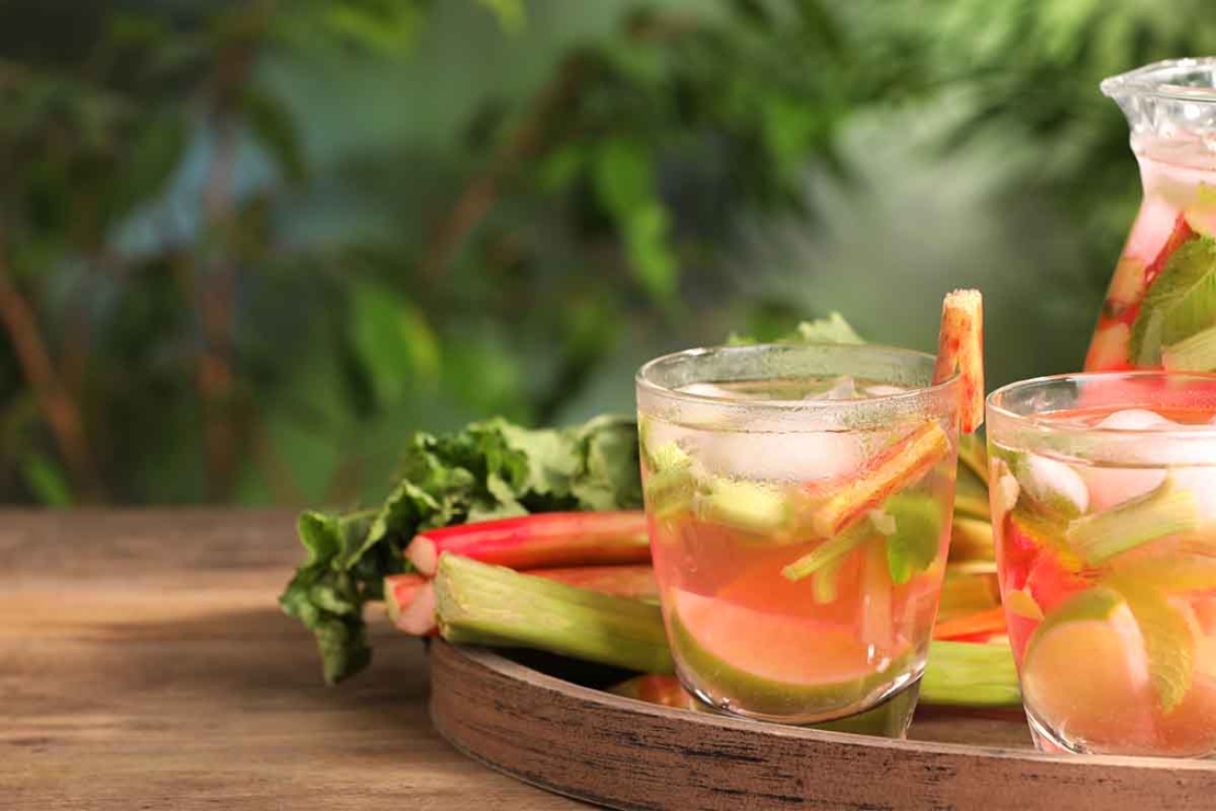Aus Rhabarber zubereitete Limonade in einem kleinen Glas, das neben Rhabarberstangen auf einem Tisch aus Holz steht, hinter dem die verschwommenen Blaetter einer Pflanze zu sehen sind. [Foto: AdobeStock_New Africa]