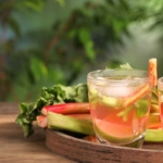 Aus Rhabarber zubereitete Limonade in einem kleinen Glas, das neben Rhabarberstangen auf einem Tisch aus Holz steht, hinter dem die verschwommenen Blaetter einer Pflanze zu sehen sind. [Foto: AdobeStock_New Africa]