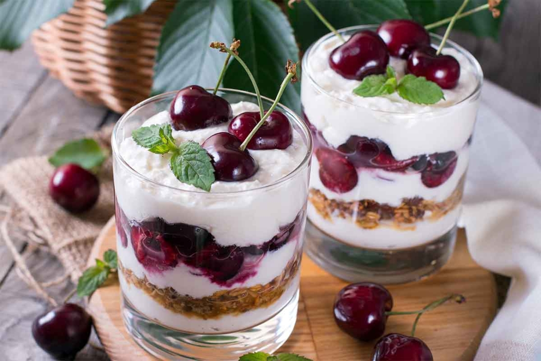 Zwei geschichtete Kirsch-Desserts stehen nebeneinander auf einem Holzbrettchen. Oben auf den Schichten aus Joghurt-Creme, Kirschen und Knusperbröseln tronen liebevoll etwas Minze und jeweils drei Kirschen zur Zierde. [Foto: AdobeStock_qwartm]