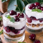 Zwei geschichtete Kirsch-Desserts stehen nebeneinander auf einem Holzbrettchen. Oben auf den Schichten aus Joghurt-Creme, Kirschen und Knusperbröseln tronen liebevoll etwas Minze und jeweils drei Kirschen zur Zierde. [Foto: AdobeStock_qwartm]