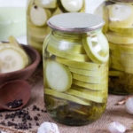 Mehrere Gläser mit eingelegten Zucchinischeiben stehen auf einem Tisch. Im Vordergrund liegt ein Holzlöffel mit Pfefferkörnern, daneben frischer Knoblauch, Dill und Schalotten. [Foto: AdobeStock_qwartm]