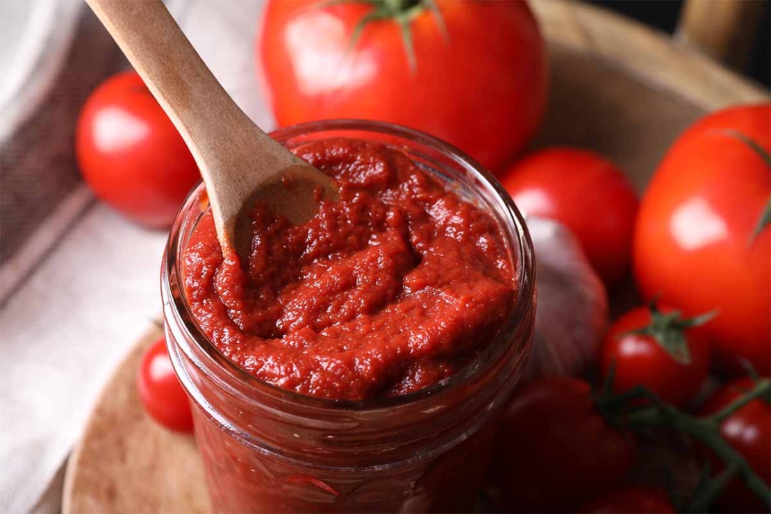 Tomatenmark mit einem Löffel in einem Glas. Im Hintergrund sind Tomaten zu sehen. [Foto: AdobeStock–New Africa]