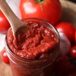 Tomatenmark mit einem Löffel in einem Glas. Im Hintergrund sind Tomaten zu sehen. [Foto: AdobeStock–New Africa]